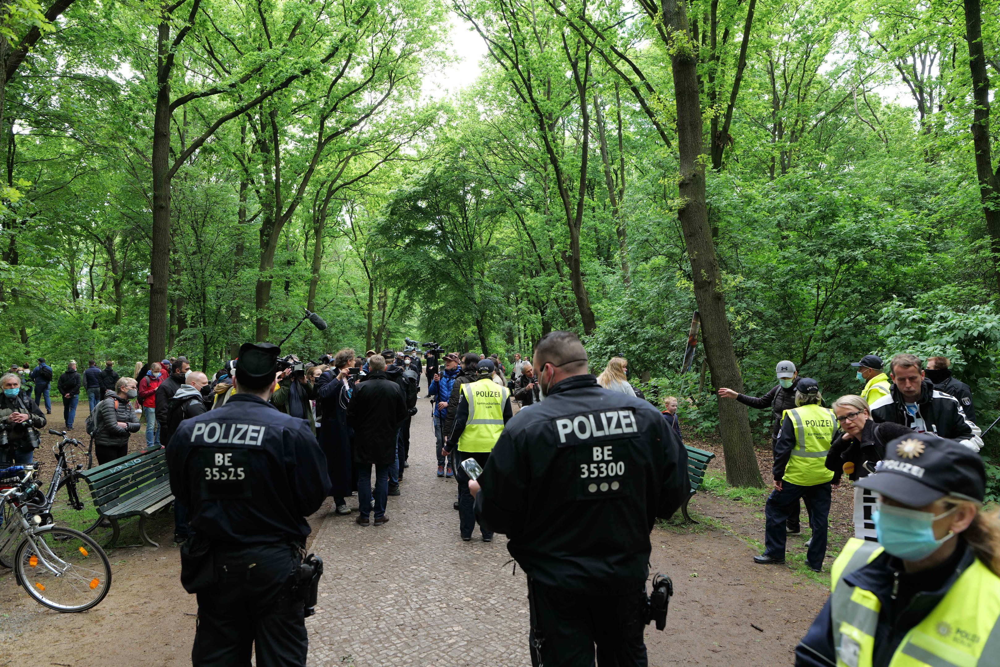 Polizeibeamte vor einer Menge während einer Anti-Terror-Demonstration in Berlin, mit Fahrrädern und einer Bank im Vordergrund und Bäumen im Hintergrund.