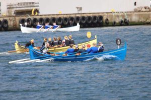 Eine Gruppe von Menschen in einem blauen und gelben Boot auf dem Wasser, die Paddel halten, mit einer Wand aus Reifen und einem Geb"ude im Hintergrund, was eine Regatta vermuten l"asst.