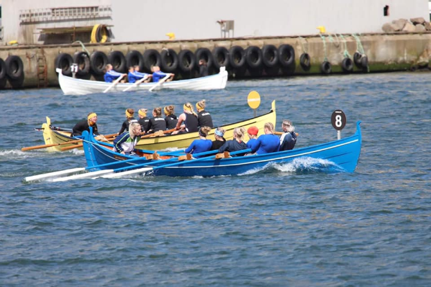 Eine Gruppe von Menschen in einem blauen und gelben Boot auf dem Wasser, die Paddel halten, mit einer Wand aus Reifen und einem Geb"ude im Hintergrund, was eine Regatta vermuten l"asst.