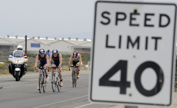 Gruppe von Radfahrern mit Helmen, die auf einer Straße mit einem Geschwindigkeitsbegrenzungsschild fahren, mit Gebäuden und Bäumen im Hintergrund unter einem klaren blauen Himmel.