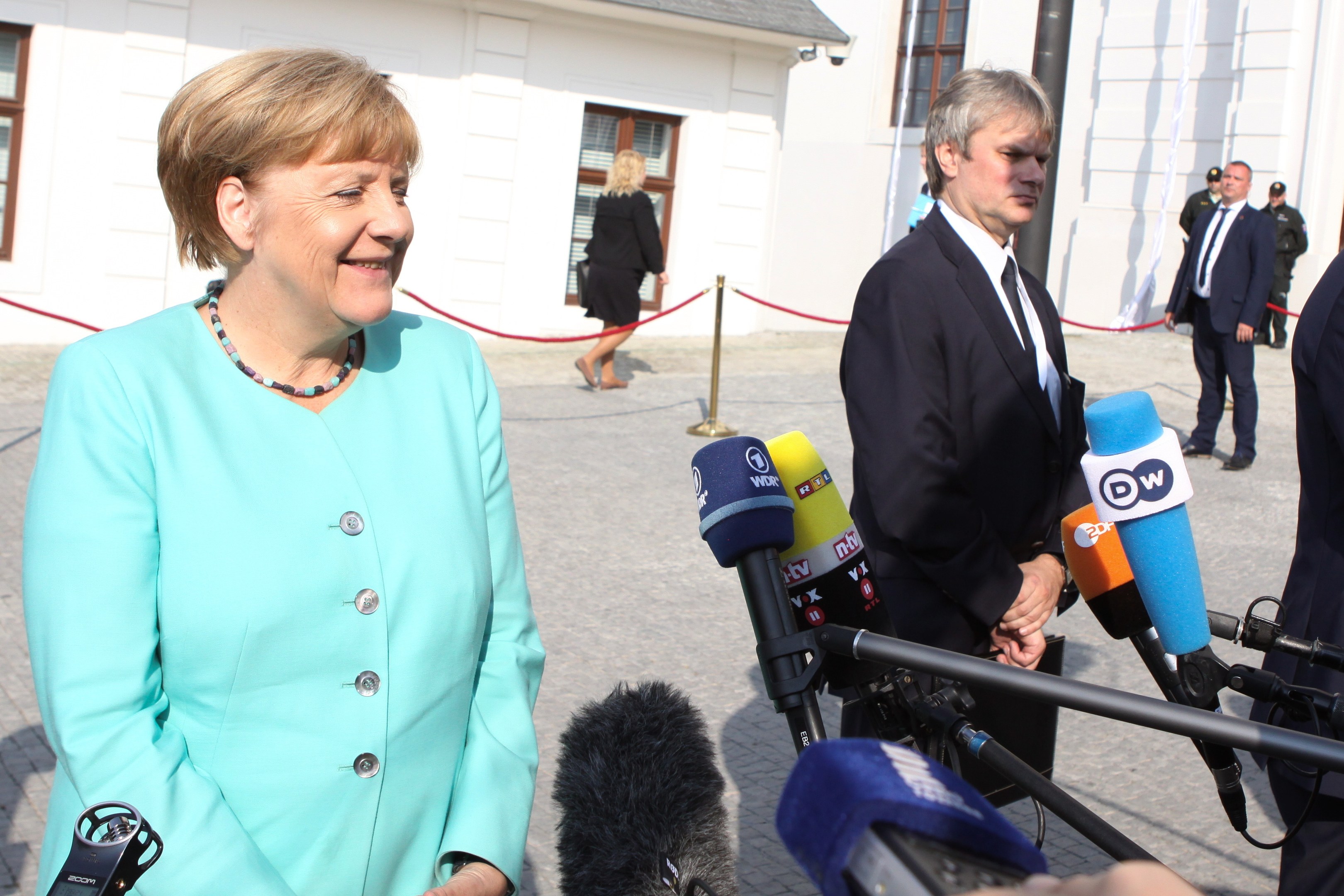 Bundeskanzlerin Angela Merkel spricht vor einem Gebäude mit Fenstern zu Reportern, umgeben von Menschen und Medienpersonal mit Mikrofonen und Ständern.