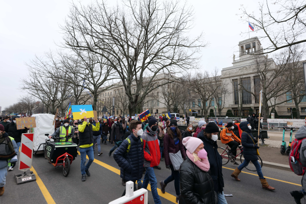 Eine große Gruppe von Menschen nimmt an einer Protestdemo auf einer Straße in Washington, D.C. teil, wobei einige Schilder und Banner halten, andere Fahrräder fahren und Schilder, Bäume und ein klarer blauer Himmel im Hintergrund zu sehen sind.