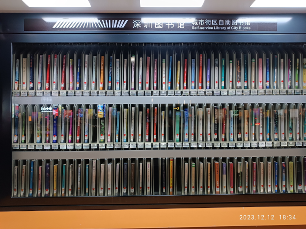 Eine gut beleuchtete Bibliothek mit hohen Bücherregalen, die mit Büchern gefüllt sind, ein sichtbares Informationsbrett und ein Wasserzeichen unten, das einer Selbstbedienungsbibliothek in einer städtischen Umgebung ähnelt.