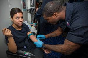 Eine Frau sitzt in einem Stuhl, während ein Mann neben ihr steht und eine Spritze hält, die darauf hindeutet, dass sie geimpft wird, mit Büchern und anderen Gegenständen auf dem Tisch vor ihnen und ein paar anderen Menschen im Hintergrund.