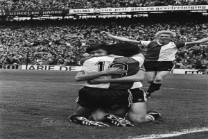 Drei Fußballspieler umarmen sich auf dem Boden und feiern ein Tor, mit jubelnder Menge und schwenkenden Fahnen im Hintergrund, dargestellt in Schwarz-Weiß.