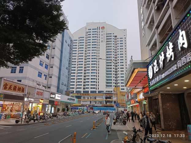 Eine belebte Stadtstraße mit hohen Gebäuden, Fahrzeugen, Fußgängern, Verkehrszeichen, Straßenmöblierung, Bäumen und einem Hotel in der Mitte unter einem klaren blauen Himmel.