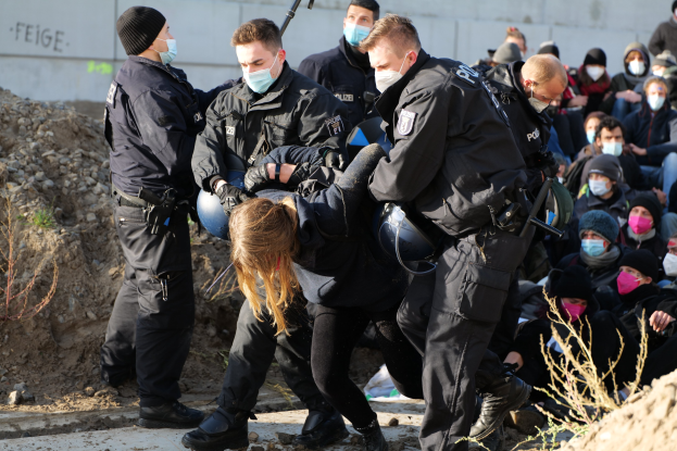 Eine Gruppe maskierter Personen in Mützen, einige bewaffnet, umgibt eine Frau, die von der Polizei eskortiert wird, mit Pflanzen, Steinen und einer protestbezogenen Wandtext im Hintergrund.