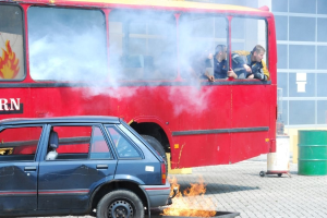 Ein roter Doppeldeckerbus mit Rauch aus dem Auspuff und einem daneben geparkten Auto, drei sichtbare Passagiere im Inneren und ein Gebäude mit Glasfenstern und einem Fass im Hintergrund.