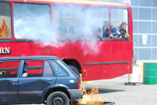 Ein roter Doppeldeckerbus mit Rauch aus dem Auspuff und einem daneben geparkten Auto, drei sichtbare Passagiere im Inneren und ein Gebäude mit Glasfenstern und einem Fass im Hintergrund.