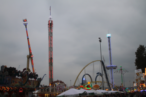 Eine nächtliche Faschingszene mit einer Menschenmenge, die sich um beleuchtete Fahrgeschäfte, Pfähle und Bäume versammelt hat.