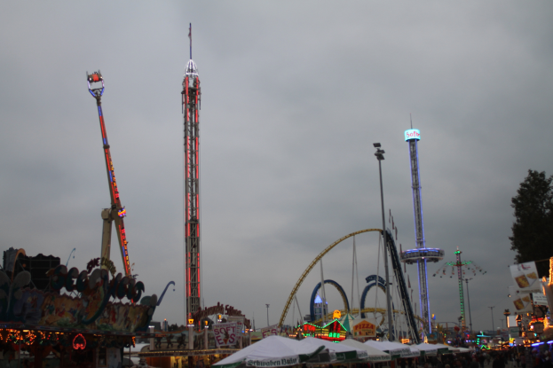 Eine nächtliche Faschingszene mit einer Menschenmenge, die sich um beleuchtete Fahrgeschäfte, Pfähle und Bäume versammelt hat.