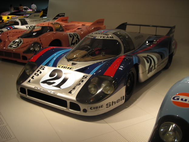 Ein Porsche 917K Le Mans Rennwagen in einem Museum ausgestellt, umgeben von anderen Sportwagen, mit Menschen in der Nähe und einer Wand im Hintergrund; ein Blatt Papier mit Text ist links sichtbar.