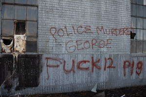 Ein Gebäude mit Graffiti, das "Polizei Mord George Fuck 12 P19" auf der Seite trägt, mit Fenstern und verschiedenen Gegenständen vorne drauf.