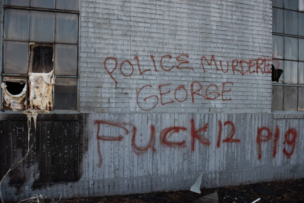 Ein Gebäude mit Graffiti, das "Polizei Mord George Fuck 12 P19" auf der Seite trägt, mit Fenstern und verschiedenen Gegenständen vorne drauf.