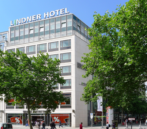 Großes Glasfrontgebäude des Lindner Hotels in Berlin, Deutschland, mit Text auf dem Dach, umgeben von Bäumen und einer belebten Straße mit Fußgängern und Fahrzeugen unter einem sichtbaren Himmel.