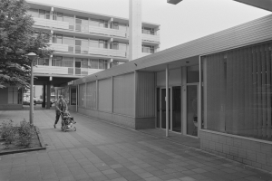 Ein Mann schiebt einen Kinderwagen über einen Gehweg in einer städtischen Umgebung, mit Gebäuden, Straßenlaternen, Fahrzeugen und Grünflächen unter einem klaren Himmel.