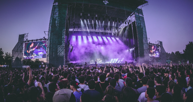 Eine große Menschenmenge bei einem Musikfestival schaut auf einer hell erleuchteten Bühne mit Bildschirmen, die vor einem Gebäude, Bäumen und einem klaren blauen Himmel steht.