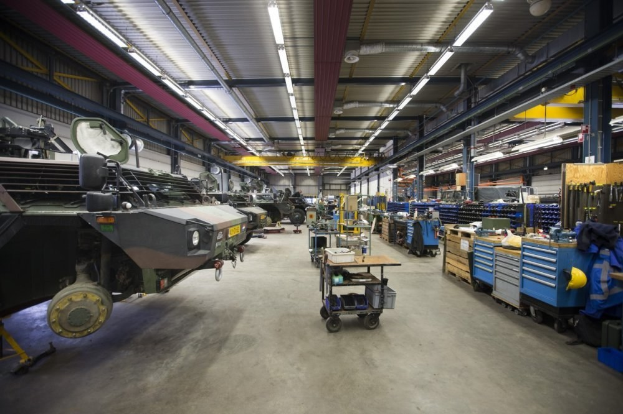 Großes Lager mit Maschinen, Fahrzeugen links, Wagen mit Gegenständen, Holzboxen und Deckenbeleuchtung.