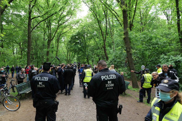 Polizisten vor einer Menge während einer Outdoor-Veranstaltung, mit Fahrrädern und einer Bank im Vordergrund und Bäumen im Hintergrund.
