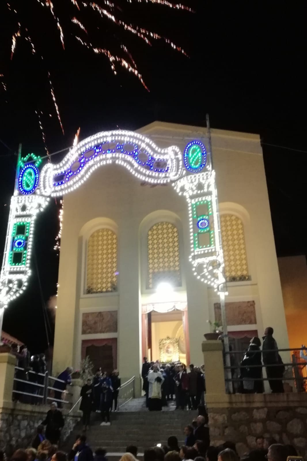 Eine Gruppe von Menschen, die vor einem Gebäude mit einem beleuchteten Bogen, Treppen mit Geländern und Topfpflanzen stehen und Silvester mit Feuerwerk im Hintergrund feiern.