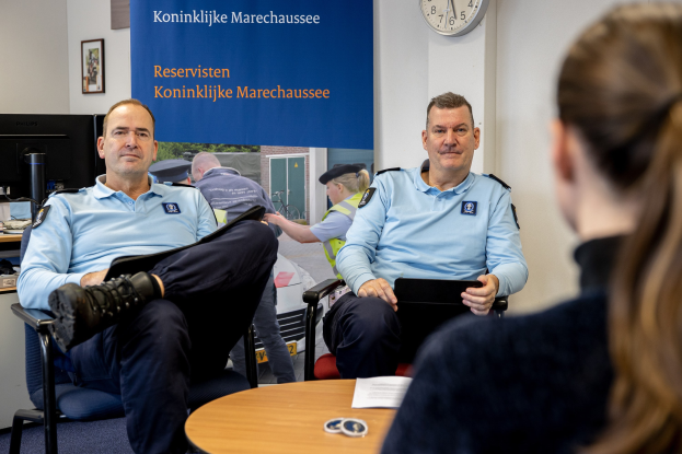 Zwei Polizeibeamte sitzen an einem Tisch mit einer Frau, Papieren und Gegenständen auf dem Tisch, einer Wandbanner und Uhr im Hintergrund, einem zweiten Tisch mit Monitor, Tastatur, Maus und Gegenständen links daneben und Fotos auf der Wand.