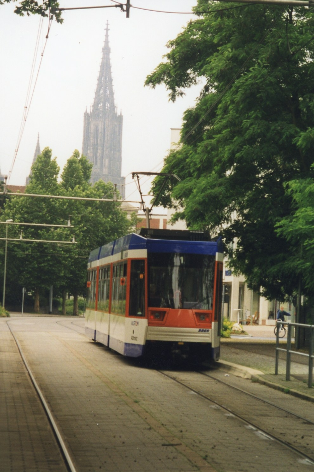 Rotes und weißes Zugfahrzeug auf Schienen neben einem hohen Gebäude, mit einer Person, die ein Fahrrad auf einem Fußweg neben den Schienen fährt, Bäume entlang der Schienen und Gebäude im Hintergrund unter einem klaren blauen Himmel.
