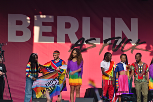 Eine Gruppe von Menschen auf einer Bühne mit einer Frau, die eine Fahne in der Mitte hält, einer Person, die eine Kamera auf einem Stativ links bedient, und einem Banner im Hintergrund mit der Aufschrift "Berlin as Fuck 2019".