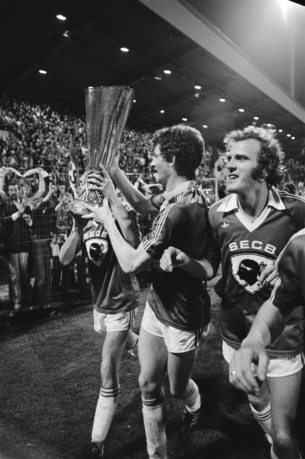 Schwarzes und weißes Bild von Fußballspielern, die einen Pokal auf einem Feld feiern, umgeben von einer Menge unter Stadionbeleuchtung.