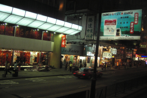 Eine nächtliche Stadtstraße mit einem fahrenden Auto, Fußgängern auf dem Gehweg, Gebäuden, Ladenschildern, Strommasten, Kabeln und Geländern bei dunkler Beleuchtung.