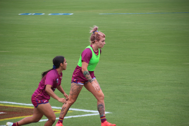 Zwei Frauen in Sportbekleidung - eine in einem grünen T-Shirt und Shorts, die andere in einem rosafarbenen Kleid - spielen Fußball auf einem Feld und zeigen sichtbare Tattoos an Händen und Füßen.