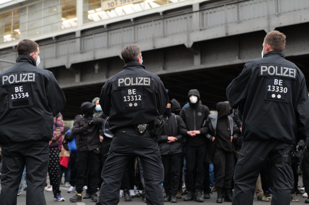 Eine Gruppe uniformierter Polizisten steht vor einer Menge von Menschen in schwarzen Uniformen und Masken, mit einer ├ťberquerung und einem Geb├Ąude im Hintergrund, w├Ąhrend einer Demonstration in einer Stadt.