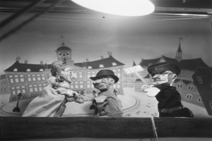 Drei Puppen in einem Raum mit einer Wand, die ein Gemälde von Gebäuden zeigt, beleuchtet von einem Licht oben auf dem Bild.