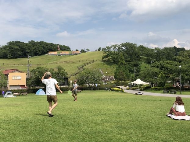Gruppe von Menschen, die Badminton in einem Park spielen, mit einem Mann, der einen Schläger hält und auf einer Decke auf dem Gras sitzt, in der Nähe von Zelten und Gebäuden, mit Hügeln und bewölktem Himmel im Hintergrund.