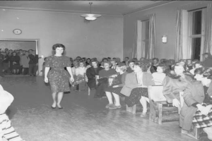 Eine Frau in einem schwarzen und weißen Kleid läuft über einen Laufsteg während einer Modenschau für Frauenrechte, mit einer Menge, die auf Stühlen sitzt und einer Wand mit einer Uhr im Hintergrund.