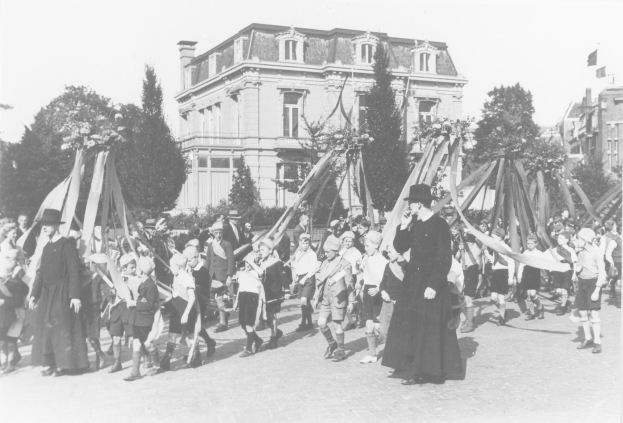 Schwarzes und weißes Bild einer Gruppe von Menschen, die eine Straße entlanggehen und Fahnen schwenken, mit Hüten, Bäumen, Gebäuden und einem Maibaum im Hintergrund.