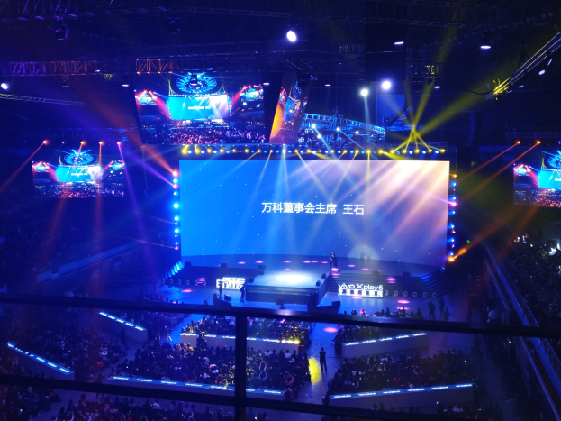 Eine große Menschenmenge sitzt vor einer hell erleuchteten Bühne mit Metallstangen und wartet auf ein E-Sport-Event, während mehrere Personen auf der Bühne stehen und ein großer Bildschirm im Hintergrund angezeigt wird.