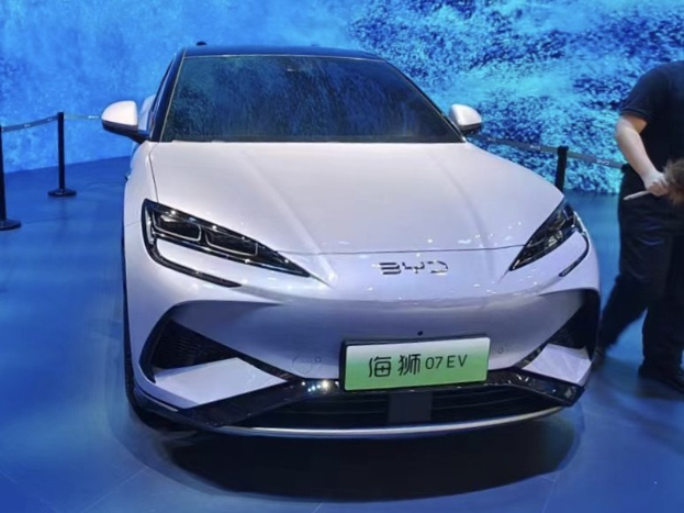 Ein weißer Toyota-Elektrofahrzeug auf einer Automesse ausgestellt, mit einer Person, die rechts daneben ein Objekt hält, Absperrpollern mit Seilen links und einem Bildschirm im Hintergrund.