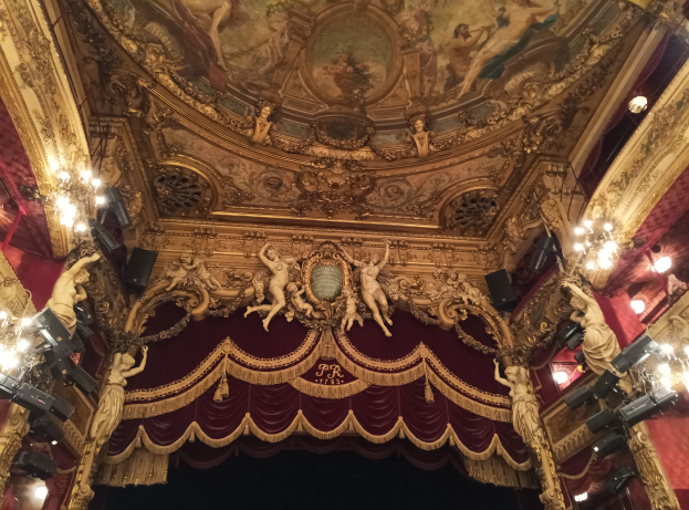 Ein Theaterinnenraum mit rotem Vorhang, Kronleuchtern, Skulpturen, Deckenmalerei und einer Bühne mit einigen Menschen davor.