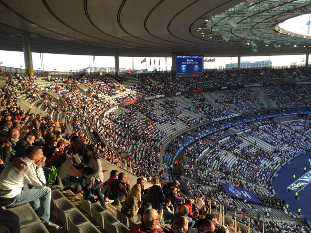 Großes Publikum in einem Stadion bei einem Fußballspiel, mit einer Bühne rechts, Fahnen, Masten, einem Bildschirm und dem Allianz Arena in München im Hintergrund.