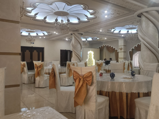 Ein großer Festsaal mit Tischen und Stühlen, die mit Tüchern drapiert und mit verschiedenen Gegenständen geschmückt sind, mit Türen und Vorhängen an den Wänden und beleuchtet von Deckenleuchten und Kronleuchtern.