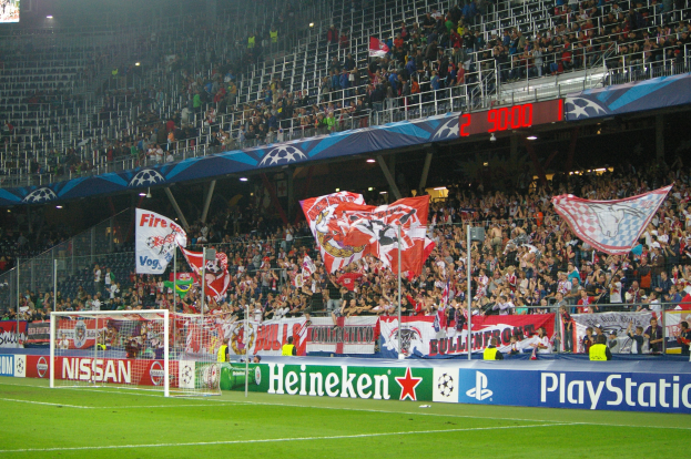 Ein Fußballspiel in einem hell erleuchteten Stadion mit einer großen Zuschauermenge, einem Tor in der Mitte, Bannern drum herum und einem Bildschirm oben links.
