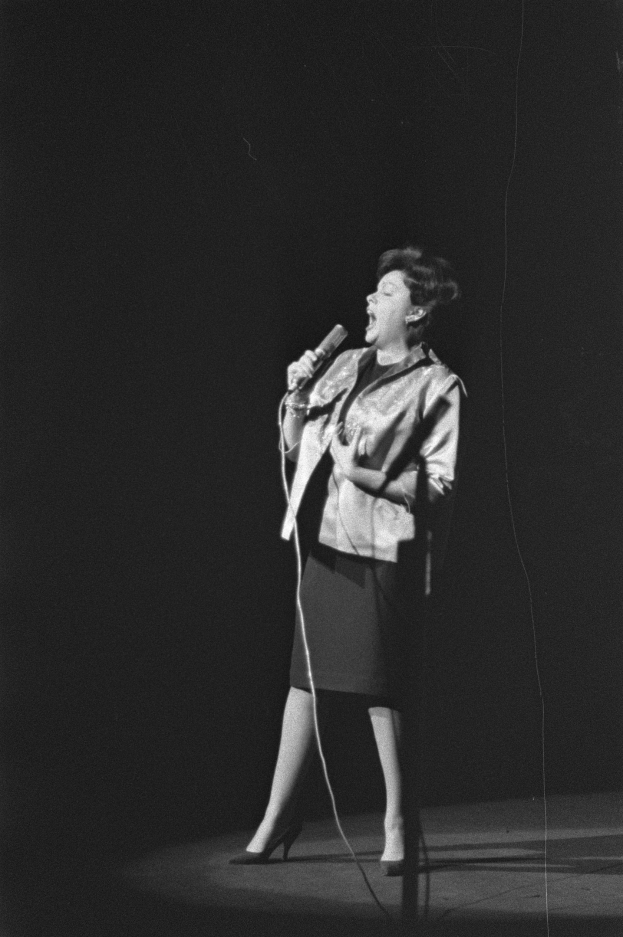 Eine Frau in einem schwarz-weißen Outfit aus den 1950er-Jahren singt fröhlich auf einer von einem Scheinwerfer beleuchteten Bühne in ein Mikrofon.