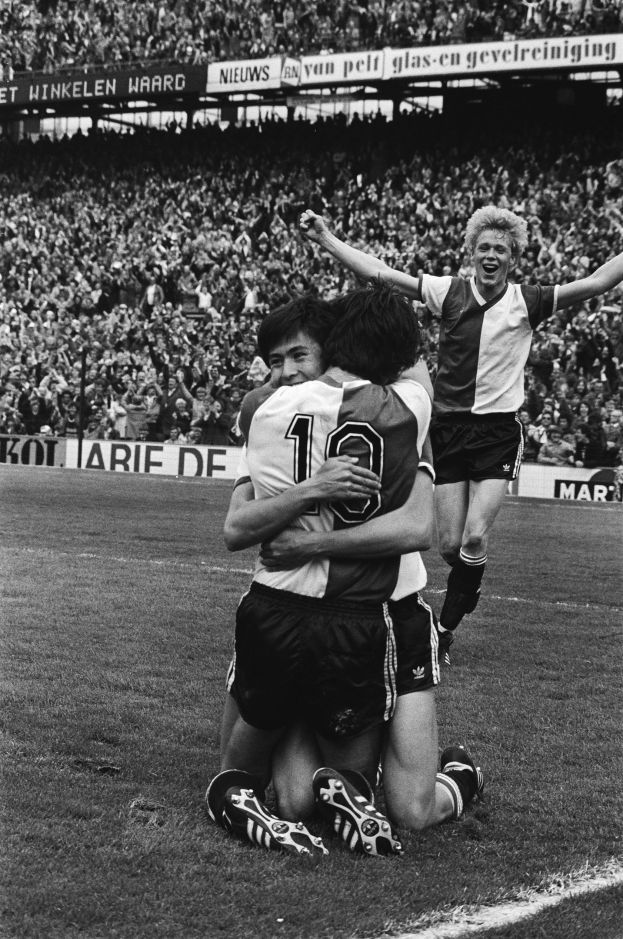 Drei Fussballspieler umarmen sich auf dem Boden nach einem Tor, mit jubelnder Menge und schwenkenden Fahnen im Hintergrund, in Schwarz-Weiß aufgenommen.