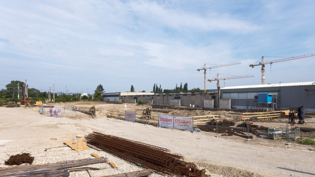 Baustelle mit verschiedenen Materialien und Ausrüstungsgegenständen, darunter Eisenstangen, hölzerne Gegenstände, bedruckte Platten, Pfähle, Drähte, Bäume, Gebäude und Kräne unter einem bewölkten Himmel.