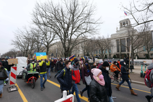 Eine große Gruppe von Menschen nimmt an einer Protestdemo auf einer Straße in Washington, D.C. teil, einige halten Schilder und Banner, andere fahren Fahrräder, und es gibt Schilder, Bäume und einen klaren blauen Himmel im Hintergrund.