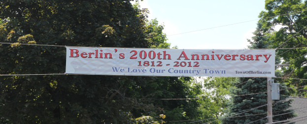 Banner mit der Aufschrift "Berliner 200. Geburtstag" hängt an einem Draht in der Mitte, Bäume im Hintergrund, ein Haus auf der rechten Seite und der Himmel oben sichtbar.