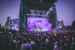Eine große Menge bei einem Musikfestival schaut Künstlern auf einer hell erleuchteten Bühne mit Instrumenten und Bildschirmen zu, die vor einem Gebäude, Bäumen und einem klaren blauen Himmel aufgebaut sind.
