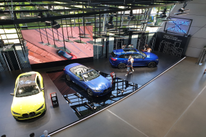 Eine Gruppe von Menschen steht innerhalb des BMW Museums in München, mit Autos zur Linken und einem Schild mit der Aufschrift "BMW Museum München" zur Rechten, überblickend eine Au├čenszene mit B├Ąumen, Geb├Ąuden und Himmel durch Glasfenster.