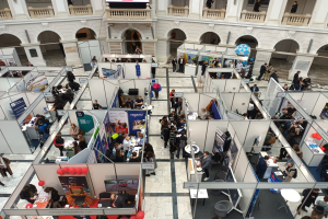 Eine große Gruppe von Menschen versammelte sich in einem Raum, der mit Ständen, Bannern, Tischen, Stühlen und Ballons gefüllt war, mit einem Gebäude im Hintergrund, das Bögen, Säulen, Geländer und einen Bildschirm aufwies.