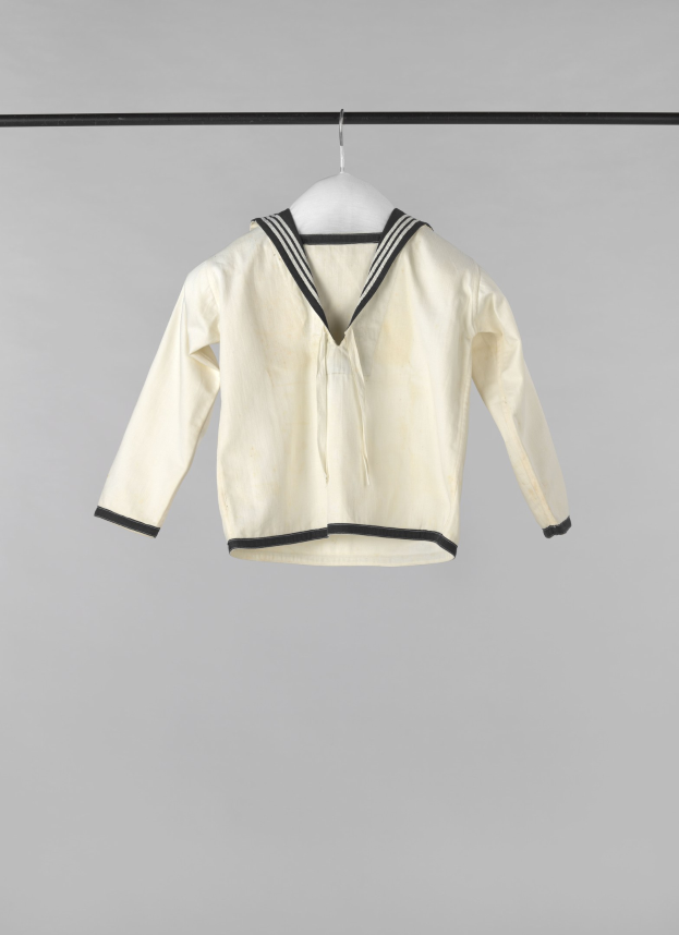 Eine weiße Matrosenjacke mit Kragen, langen Ärmeln und Knopfleiste, die an einer Wäscheleine gegen einen weißen Hintergrund hängt, aus leichtem Rippenstoff.